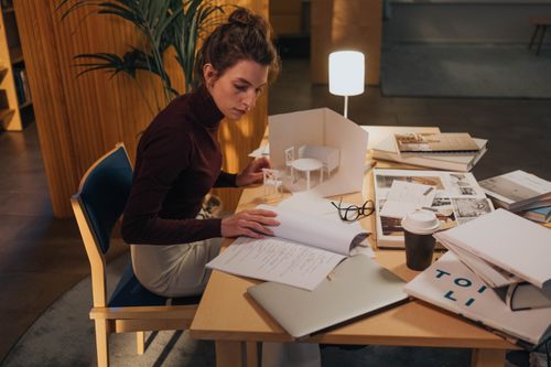 Free A young woman concentrating on architecture designs at a desk, surrounded by books and a laptop. Stock Photo