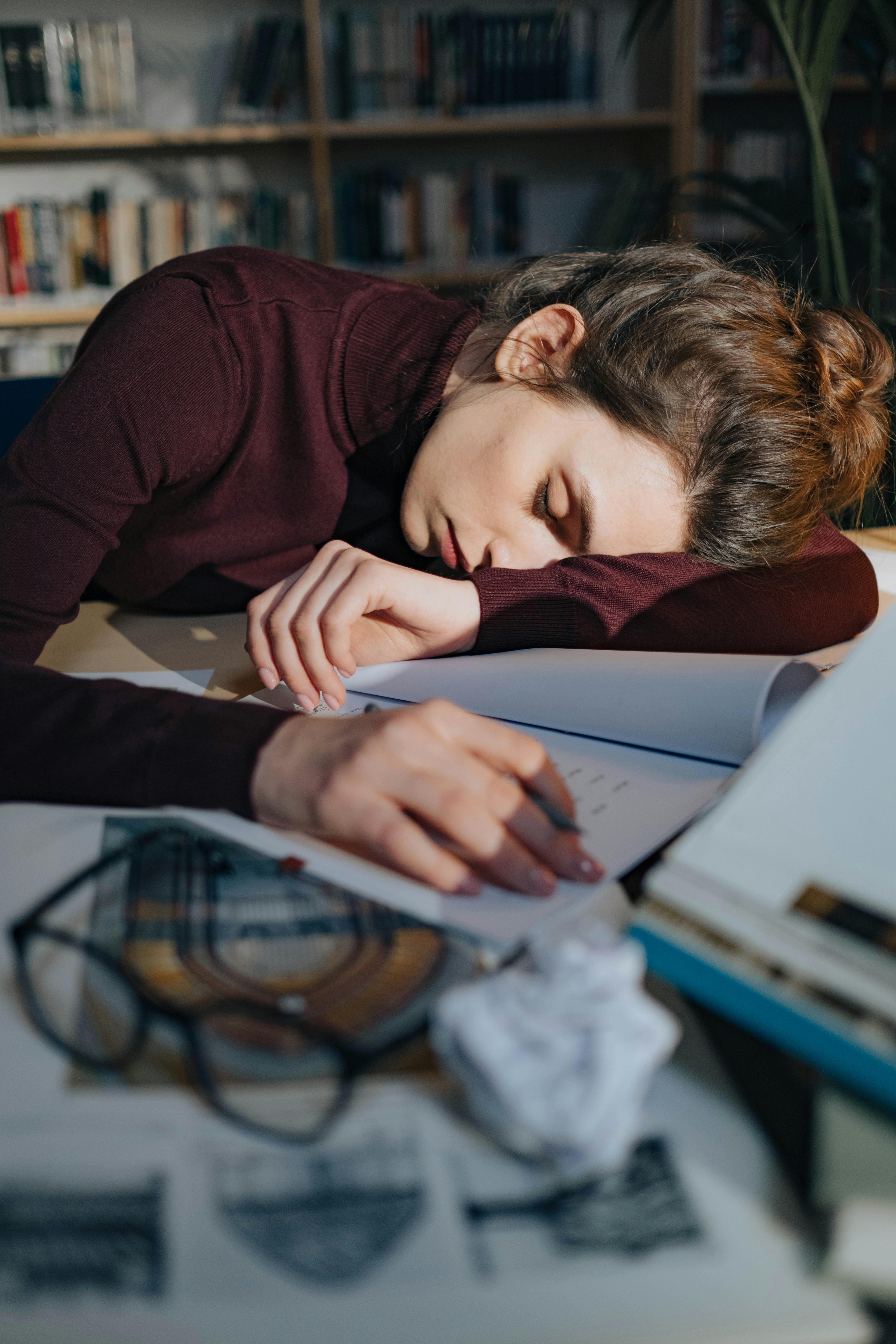 A tired woman sleeping on a desk surrounded by documents and a book.