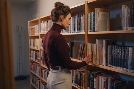 A woman browsing a bookshelf indoors, captured in a peaceful library setting.