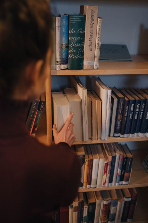 Free A woman selects a book from a tall wooden bookshelf in a library. Stock Photo