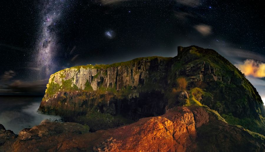 The Milky Way rises over a sea cliff lit warm by distant lights at night