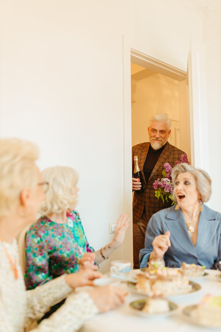 An Old Man Wearing Brown Checkered Suit Holding A Wine In A Door Near A Group Of Old Ladies