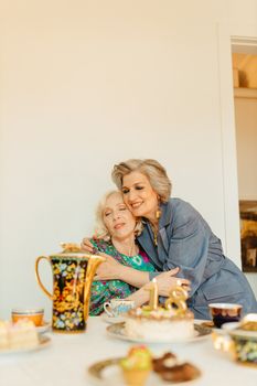 Two senior women joyfully celebrating a birthday with cake and tea indoors.