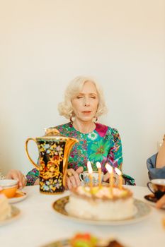 Senior woman in a colorful dress at a birthday party with cake and teapot set in a cozy indoor setting.