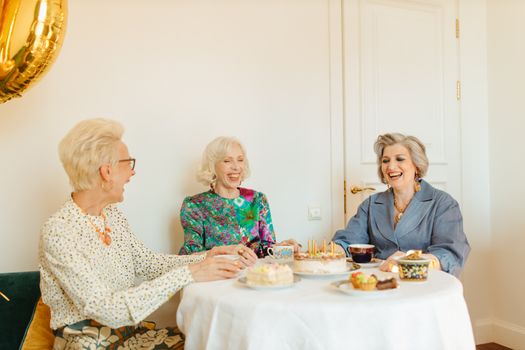 Three senior women laughing and enjoying a tea party with cakes indoors.