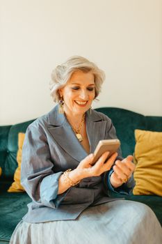 Elderly woman with gray hair enjoys using her smartphone at home, smiling while seated.