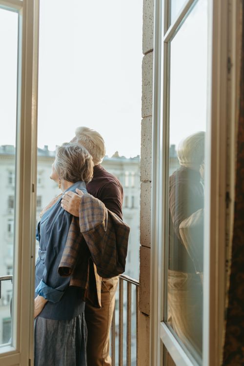 Free Senior couple standing on a balcony, embracing and enjoying the view through large glass windows. Stock Photo