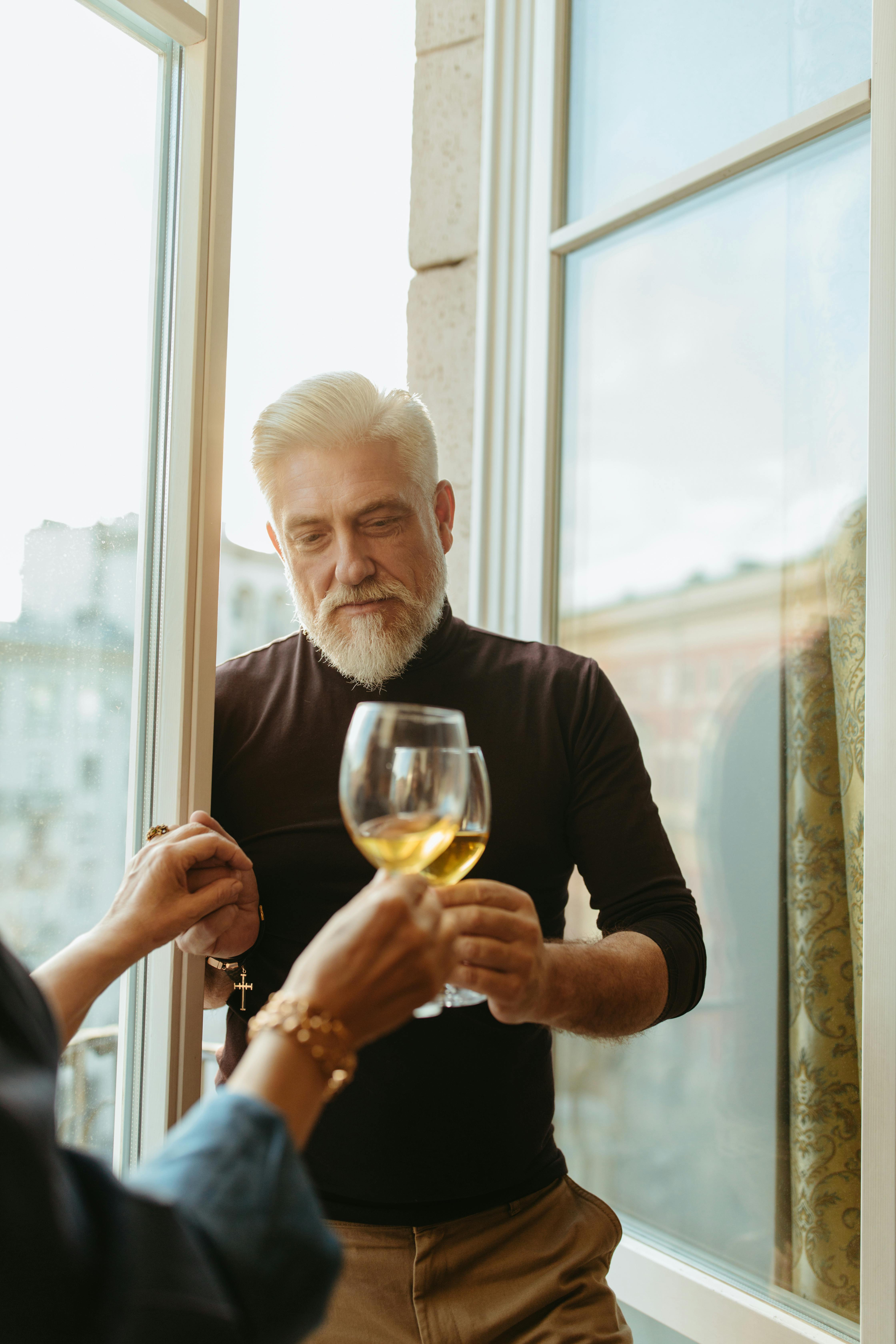 An elderly man with gray hair toasting wine indoors by a window with another person.
