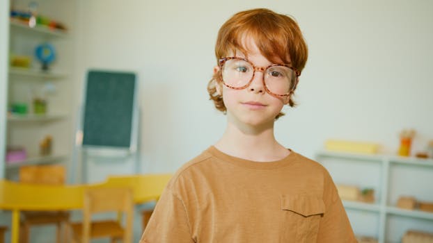 Young redheaded boy with glasses in a classroom wearing a brown shirt.