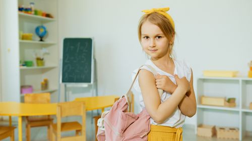 Free A young girl in a classroom holding a backpack, surrounded by educational materials. Stock Photo