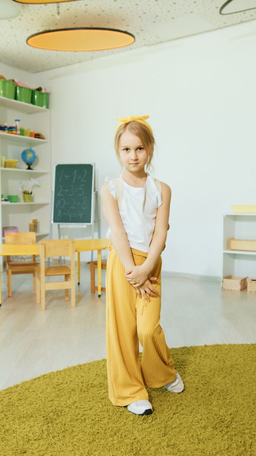 Free A child in a white top and yellow pants in a light-filled classroom with educational decor. Stock Photo