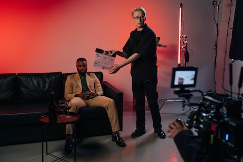 A Man in Black T-shirt Holding Holding a Clapperboard Near a Man Sitting on Black Sofa