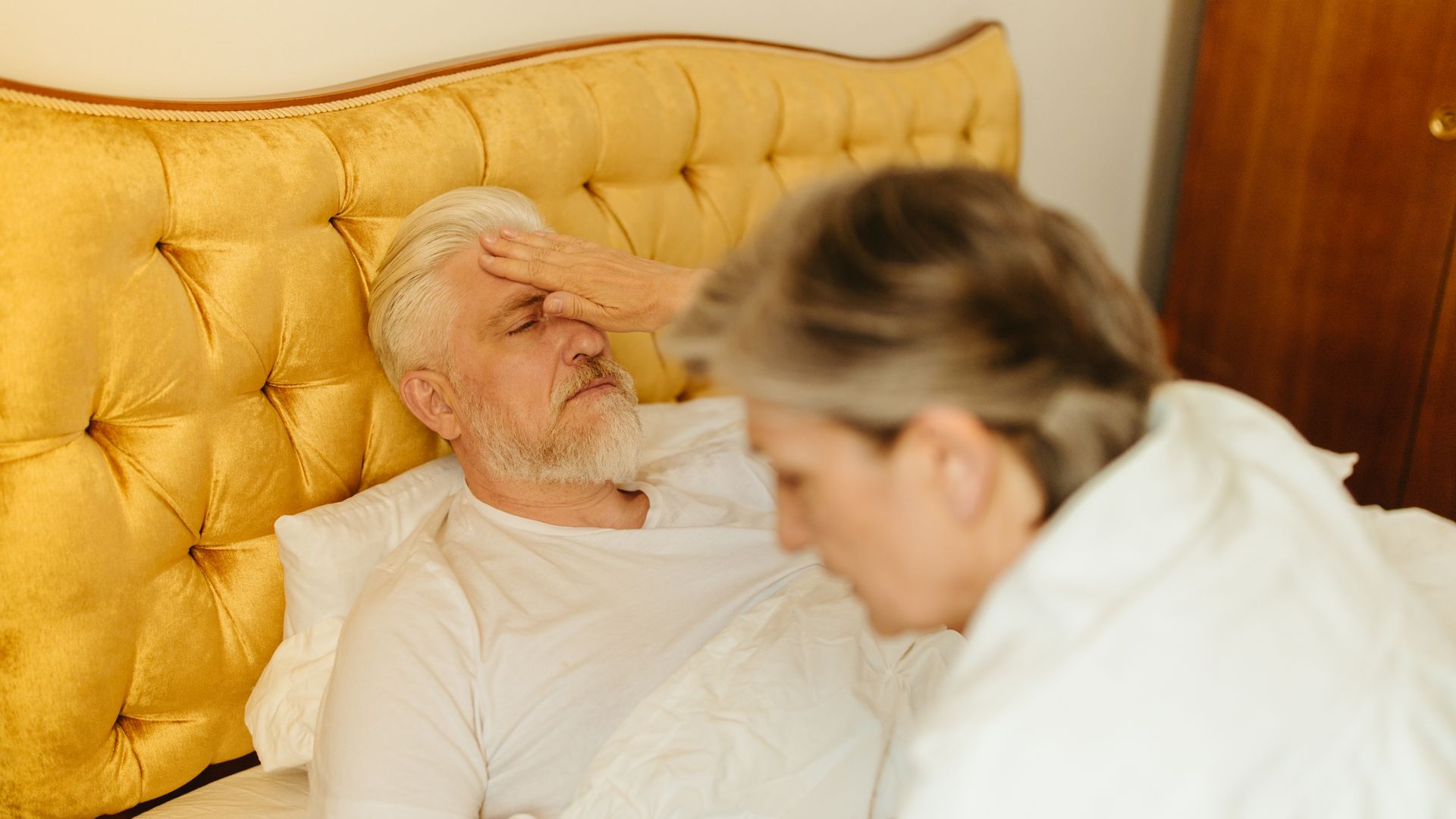 Modafinil Use in Elderly Patients: Exploring Benefits and Considerations An elderly man in bed being checked for fever by a caregiver, suggesting illness or resting state — modafinil use in elderly patients