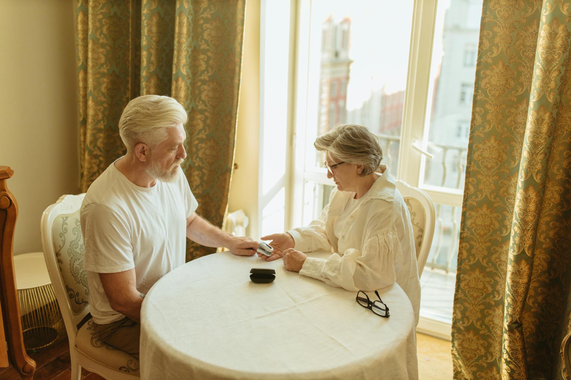 A senior couple checks their health with a finger pulse oximeter at a table in a warmly lit room.