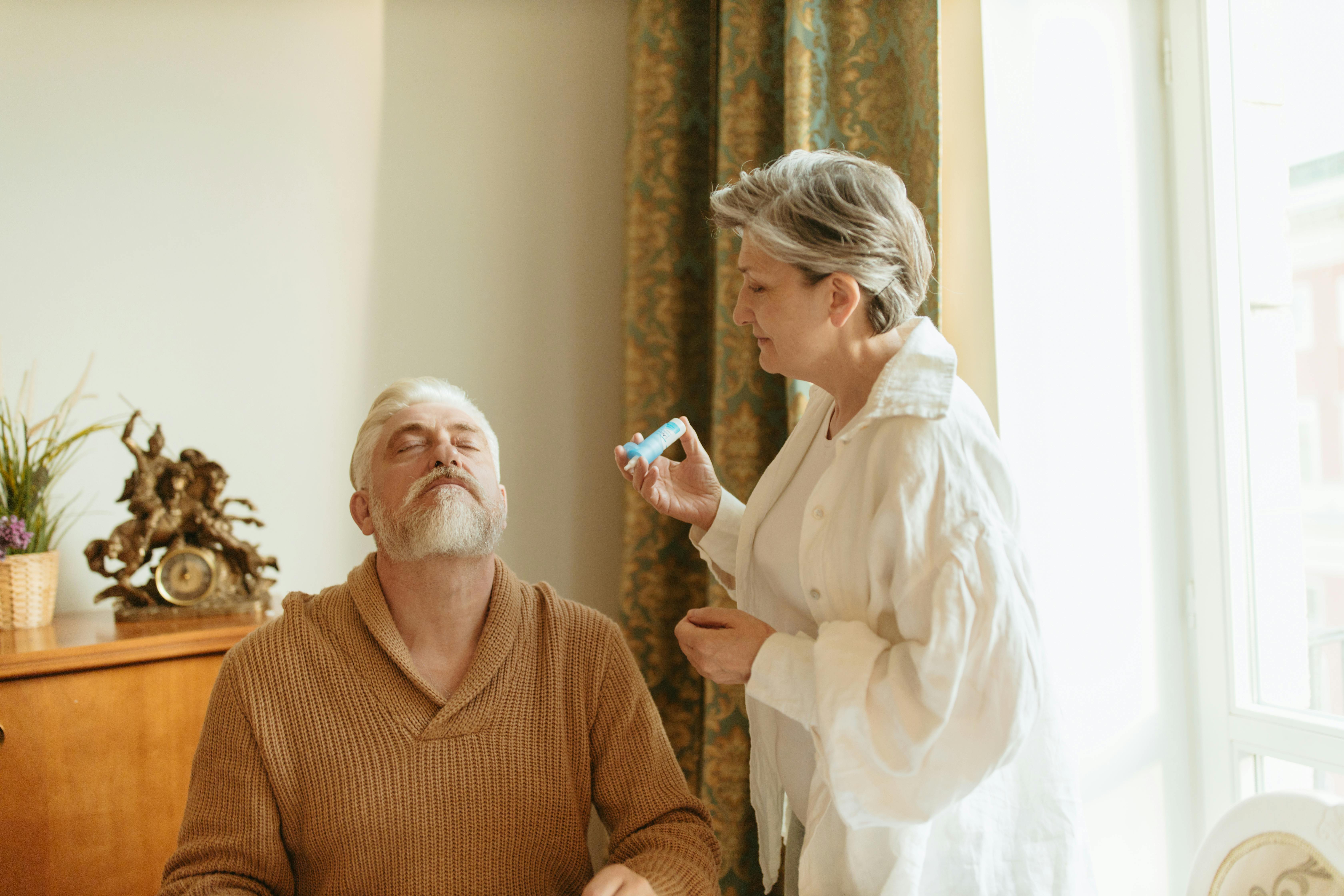 An elderly woman assists her husband with health care indoors by the window.