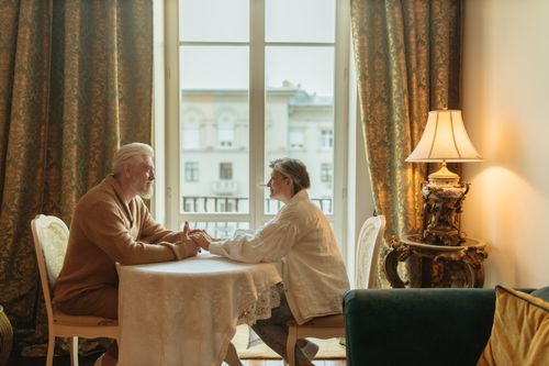 Free An elderly couple holding hands and smiling at each other in a cozy indoor setting with elegant decor. Stock Photo