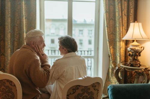 Free An elderly couple shares a tender moment by the window in a warmly lit room. Stock Photo