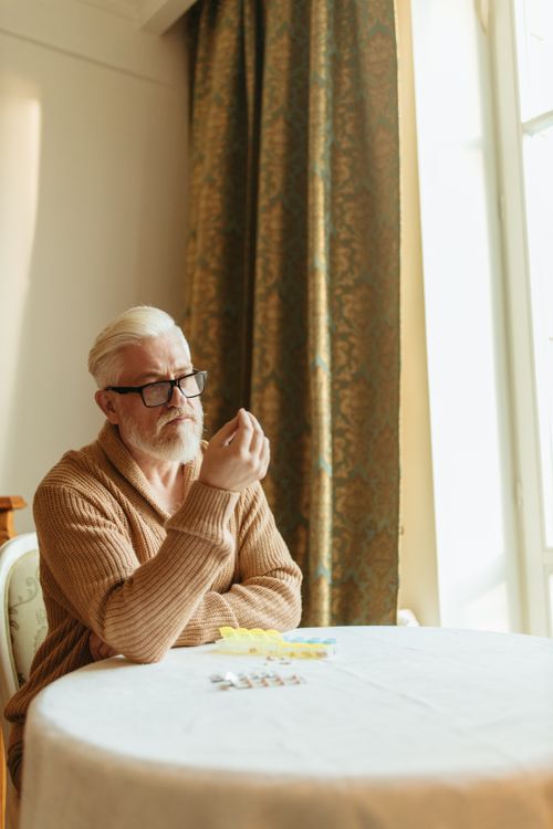 Free Elderly man with white hair and glasses contemplating medication at a sunlit indoor table. Stock Photo