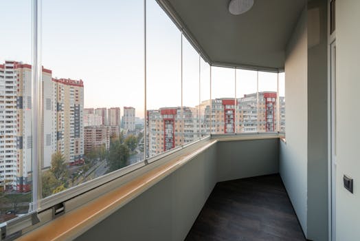A modern balcony with glass panels overlooking a city's high-rise buildings at sunset.