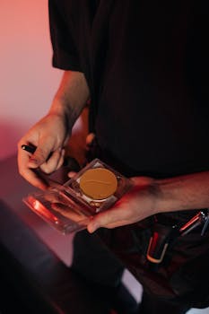 A makeup artist prepares foundation with tools in hand, under warm lighting.
