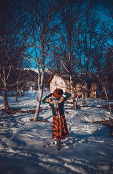 Back view of anonymous girl in warm jacket and skirt with hat standing on snowy ground among leafless trees near aged building under blue sky