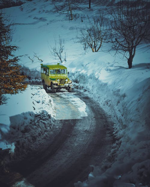 Yellow aged automobile driving on narrow curvy roadway along snowy terrain with leafless trees