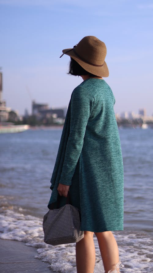 Woman In Green Coat With Brown Hat Standing On Seashore With A Handbag