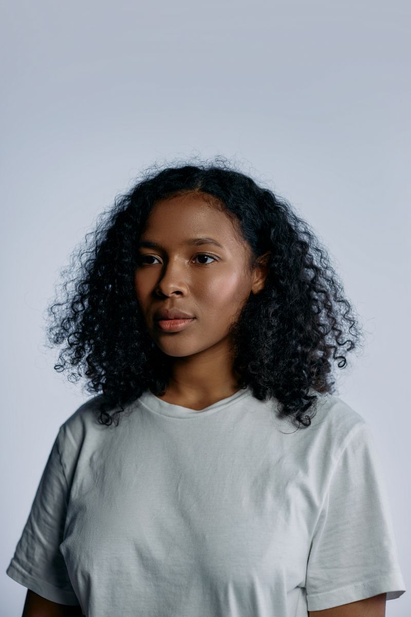 Woman with curly hair in a pale t-shirt against a soft grey studio wall — photo by cottonbro studio on Pexels