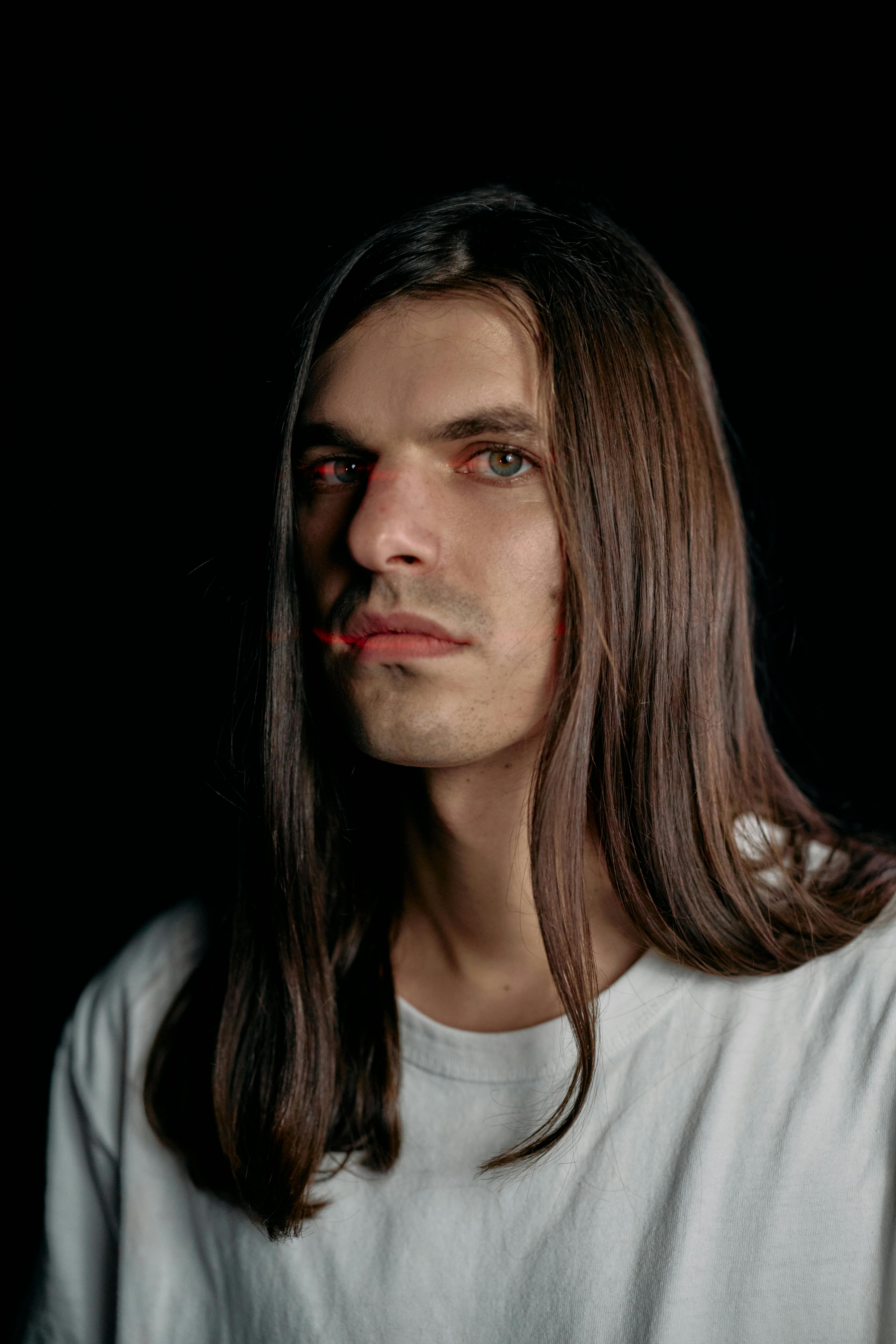 A medium close-up portrait of a man with long hair against a black background, showcasing a neutral expression.