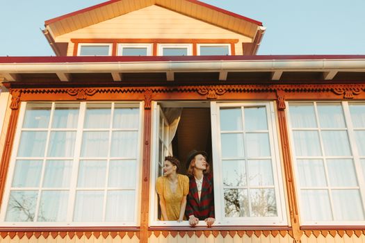 A couple in vintage attire leans out of a picturesque window under a clear sky.
