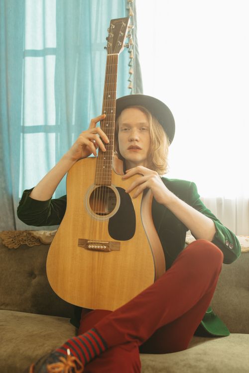 Free A young musician posing with an acoustic guitar on a sofa in a warm, indoor setting. Stock Photo