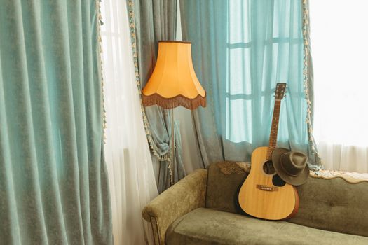 Vintage living room with acoustic guitar, hat, and retro lamp by a window with curtains.