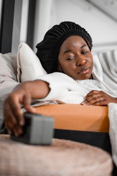 Black woman lying in bed reaching to turn off an alarm clock.