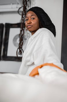 A young woman wearing a hair cap and white hoodie sits on bed indoors, serious expression.