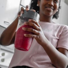 Smiling woman holding a personal blender cup of red smoothie with a grey blade base, in a white kitchen