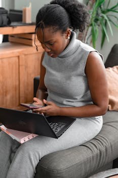 A woman multitasks with a smartphone and laptop while sitting comfortably on a couch at home.
