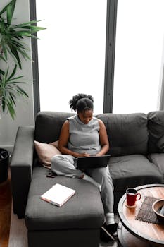 African American woman sitting on couch working remotely on laptop in a cozy home setting.