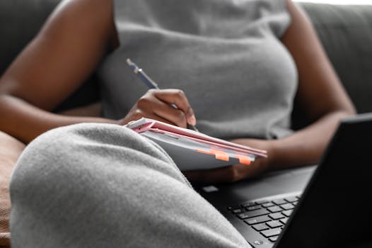 Woman concentrating on writing in a notebook while using a laptop indoors.