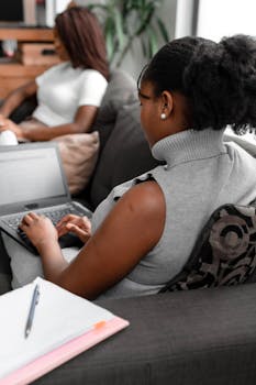 Two women using laptops in a cozy living room setting, collaborating from home.
