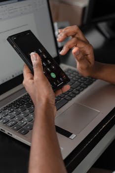 Close-up of hands using a smartphone while working on a laptop, showcasing multitasking skills.