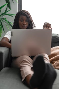 African American woman working remotely on a laptop at home, relaxing on a comfortable couch.