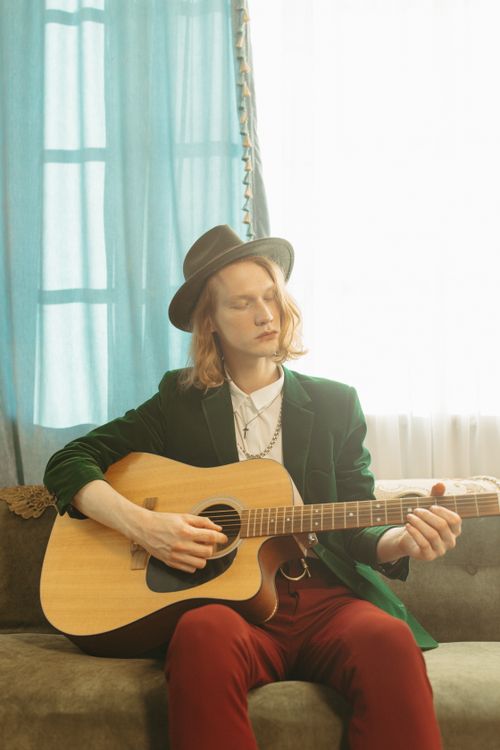 Free A young adult musician with long hair playing an acoustic guitar while sitting on a couch indoors. Stock Photo