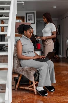 Two African American women collaborating indoors, using laptops while sitting and standing.