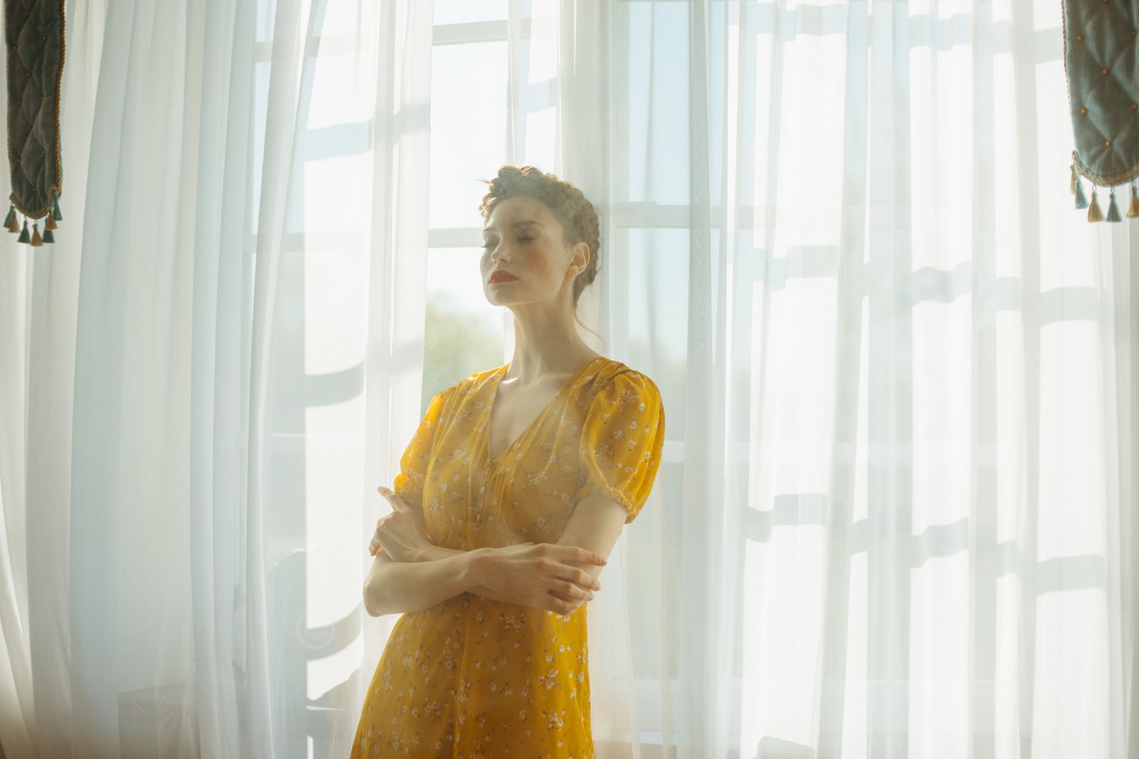 Woman in a yellow floral dress stands with arms crossed beside sunlit sheer white curtains