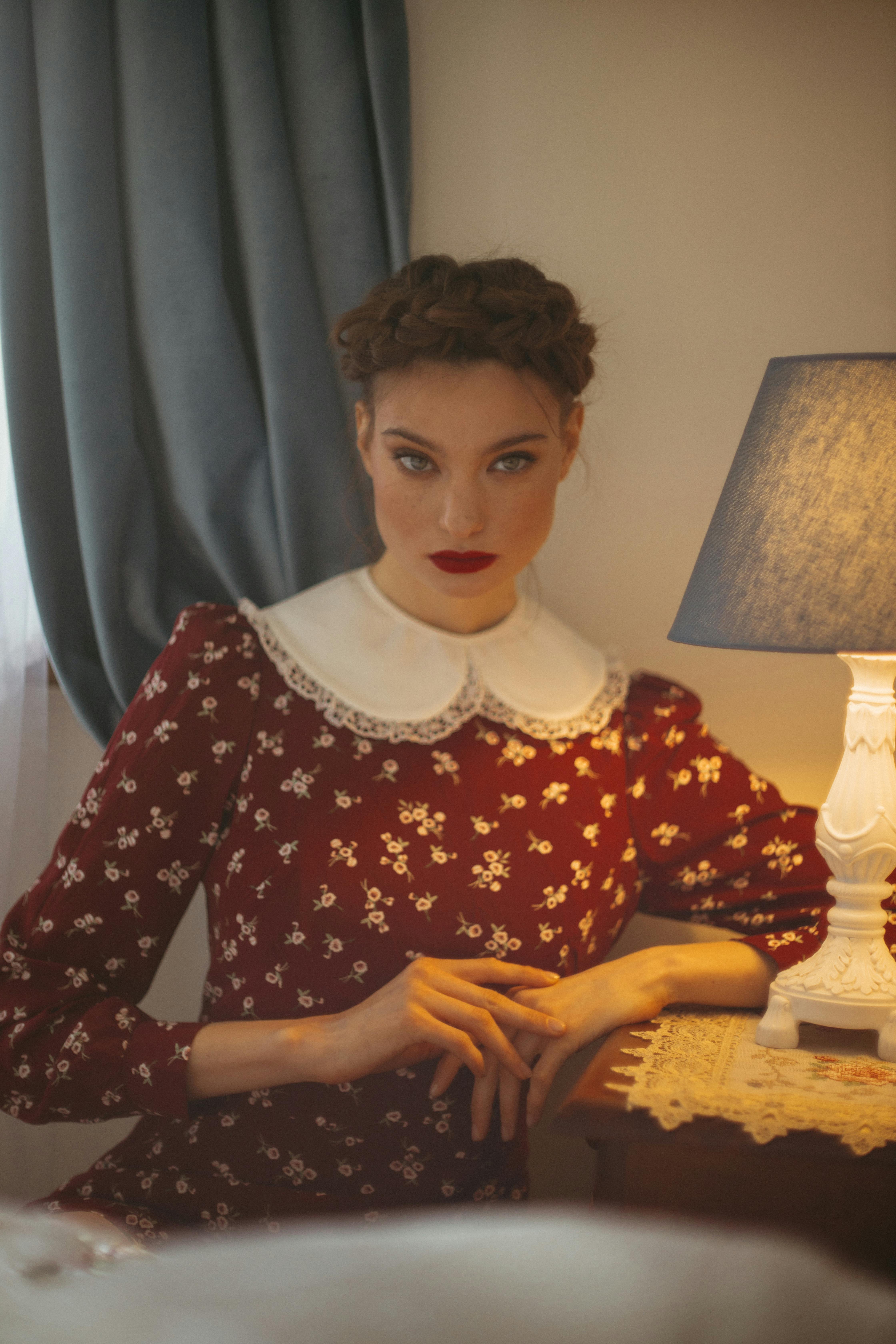 Portrait of a woman with braided hair and red lips, wearing a vintage floral dress beside a lamp.