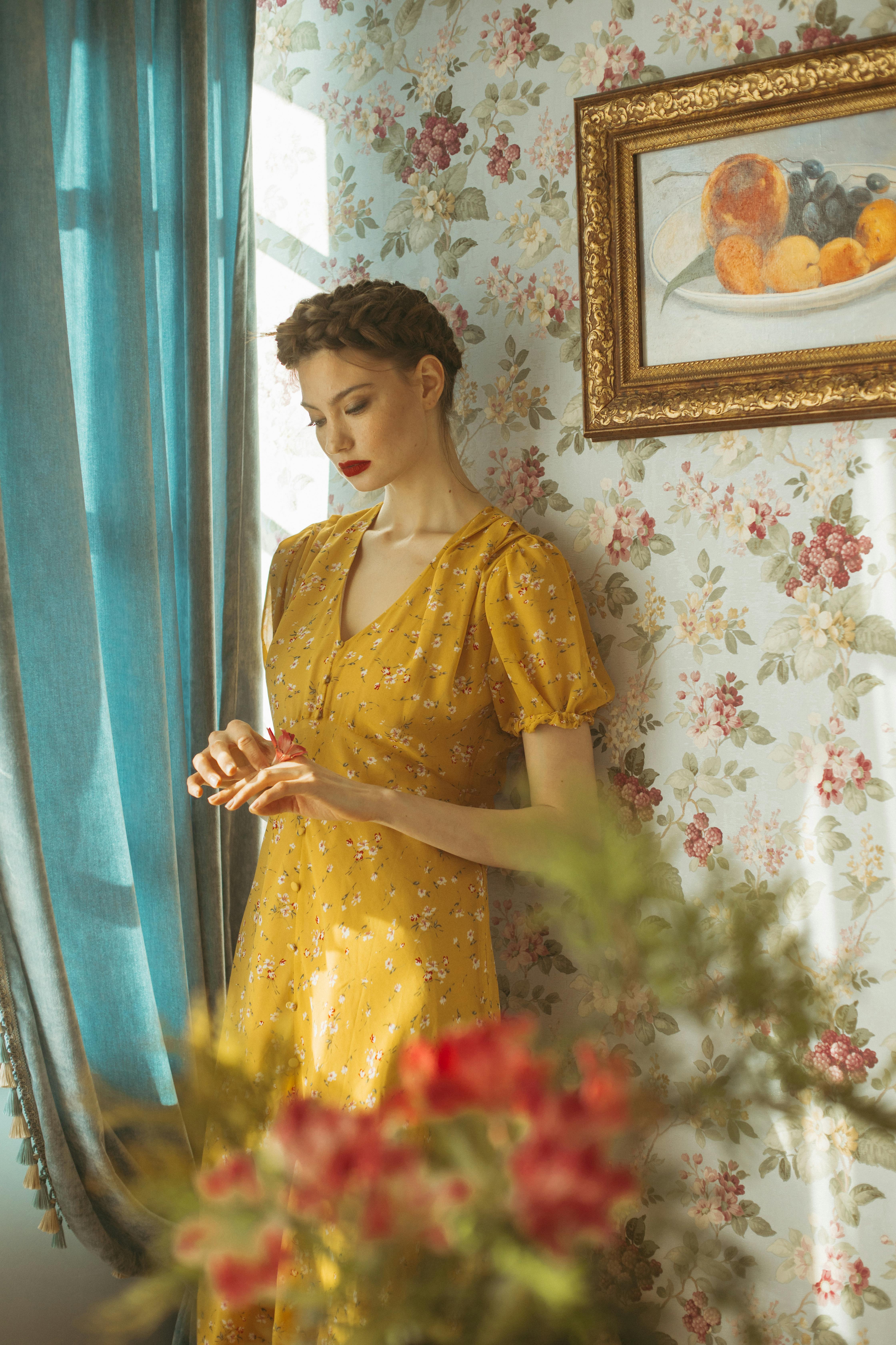 A contemplative woman in a yellow dress stands by a sunlit window in a flower-themed room.