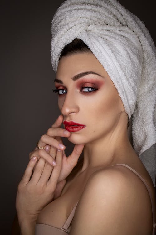 Stylish female model with evening makeup and towel on wet hair standing on gray background and looking at camera with confident seductive look