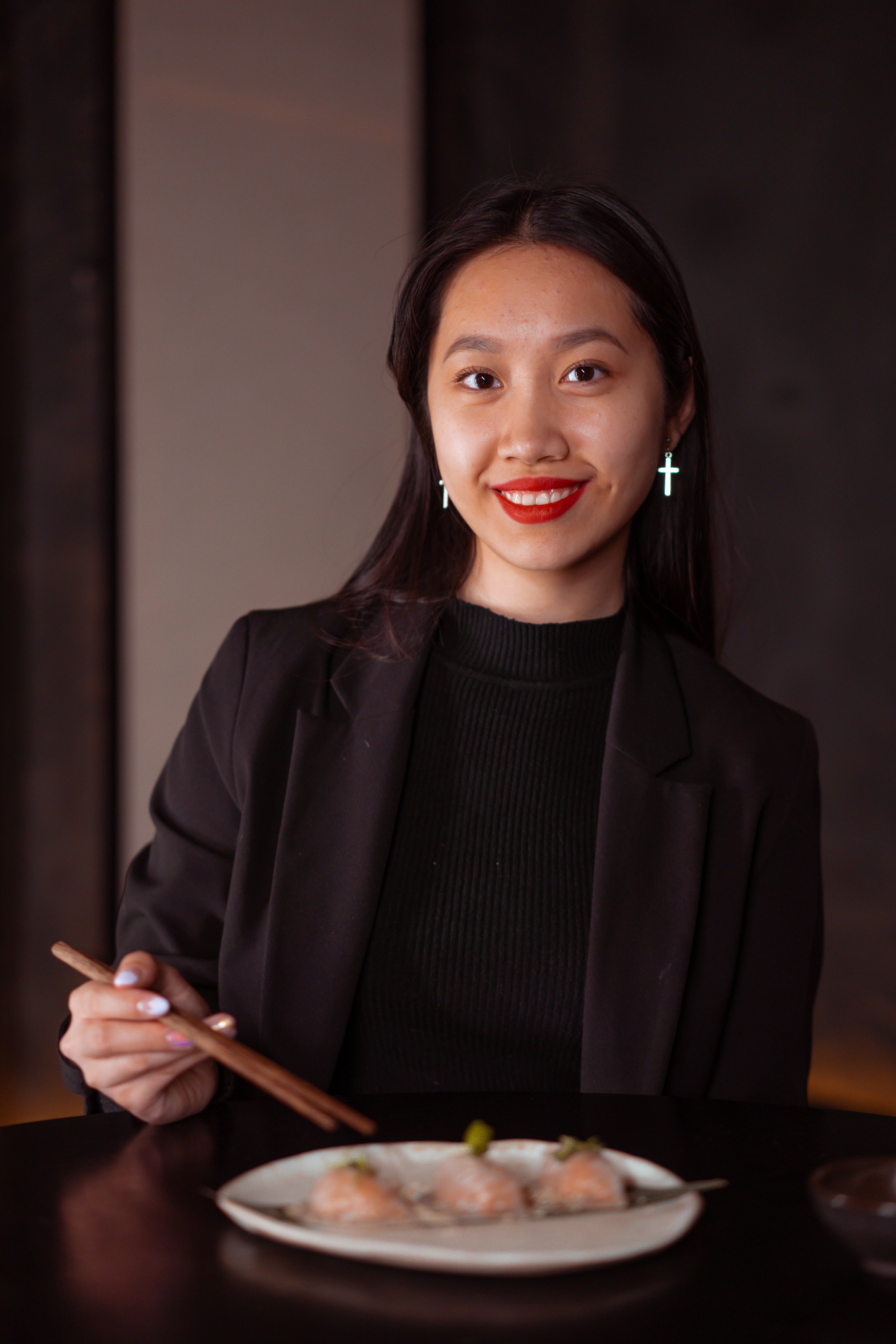 Asian woman in a restaurant enjoying dumplings with chopsticks, smiling at the camera.
