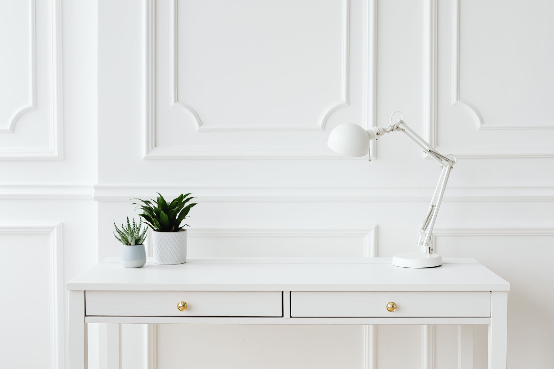 A minimalist white desk setup featuring potted plants and a sleek desk lamp against a white wall.