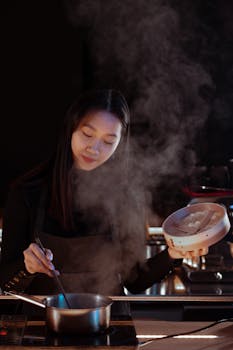 Asian woman cooking in a kitchen with visible steam, focused and serene.